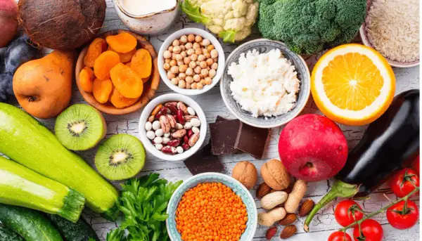 Kolorowy stół pełen zdrowych produktów: cukinia, kiwi, pomidory, bakłażan, jabłko, pomarańcza, brokuły, kalafior, soczewica, ciecierzyca, fasola, orzechy, migdały, suszone morele, ryż, twaróg i gorzka czekolada – idealne warzywa o niskim indeksie glike...