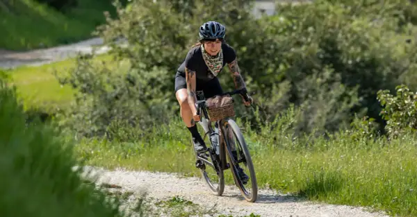 Kobieta z tatuażami na rowerze, ciesząca się kolarstwem gravelowym na piaszczystej drodze wśród zieleni.