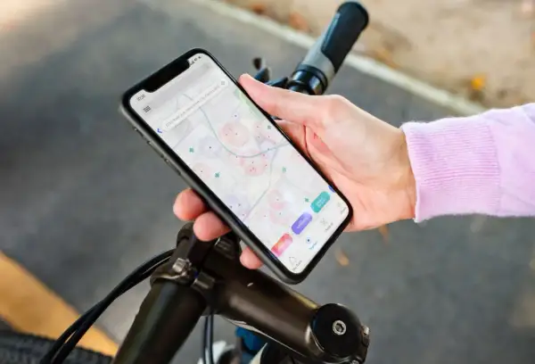 Dłoń trzyma smartfon z mapą i ikonami aplikacji rowerowych, oparty o kierownicę roweru.
